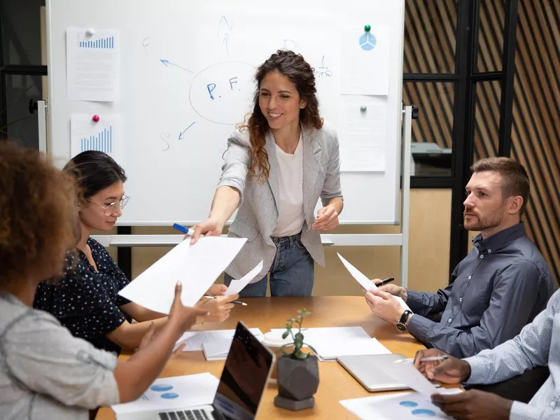 Smiling female leader give paper report to employees at meeting